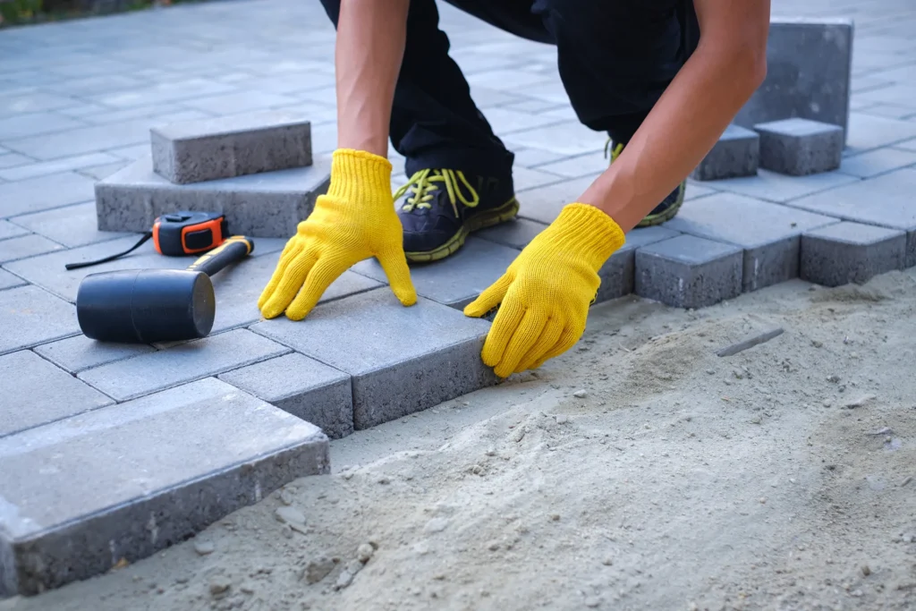 Construction worker wearing bright yellow gloves carefully setting a gray concrete paver into a sand base while installing a driveway or patio.